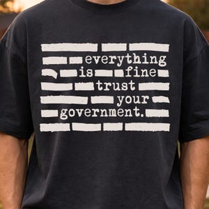 Puede incluir: Camiseta gris oscuro con el texto blanco "everything is fine trust your government". El texto está dispuesto horizontalmente, con cada palabra o frase separada por una barra horizontal blanca.
