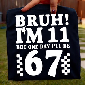 Könnte beinhalten: Schwarzes T-Shirt mit weißem Text: "BRUH! I'M 11 BUT ONE DAY I'LL BE 67". Die Zahl 67 hat ein kariertes Flaggenmuster auf beiden Seiten. Das T-Shirt wird von einer Hand gehalten.