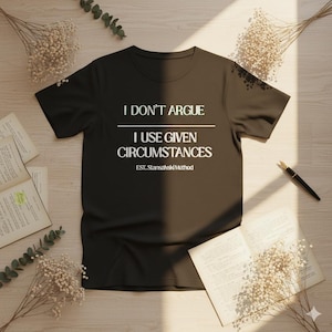 Può includere: Una maglietta nera con testo bianco: "I DON'T ARGUE, I USE GIVEN CIRCUMSTANCES, EST. Stansahski Method." La maglietta è su una superficie chiara, con libri aperti, fiori secchi e una penna. Un capo d'abbigliamento casual.