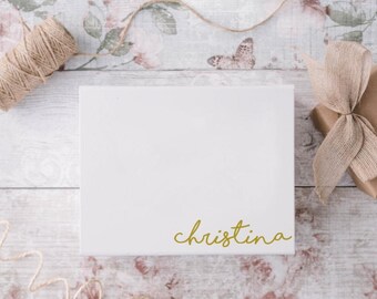 May include: A white gift box with the name "Christina" in gold script. The box is on a floral and wood-grain patterned surface, with twine and a wrapped gift with a bow.