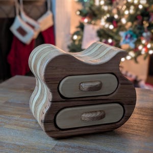 May include: A wooden jewelry box with two drawers. The box is made of light and dark wood and has a unique, wavy shape. The drawers have wooden knobs.