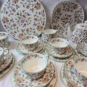 May include: A set of floral patterned teacups, saucers, and a teapot with a matching serving tray. The teacups and saucers are white with a green border and a floral pattern in shades of pink, purple, and green. The teapot is white with a green border and a floral pattern in shades of pink, purple, and green. The serving tray is white with a green border and a floral pattern in shades of pink, purple, and green.