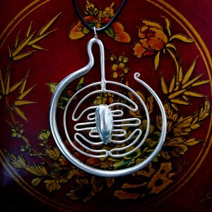 Peut inclure: Collier avec pendentif en argent avec un motif circulaire présentant une forme stylisée et abstraite au centre. Le pendentif est suspendu sur un fond rouge avec des motifs floraux dorés.
