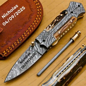 Könnte beinhalten: Ein Klappmesser aus Damaststahl mit gemusterter Klinge und Holzgriff. Auf der Klinge des Messers sind der Name "Nicholas" und das Datum "04/09/2025" eingraviert. Eine Lederscheide ist im Hintergrund zu sehen.