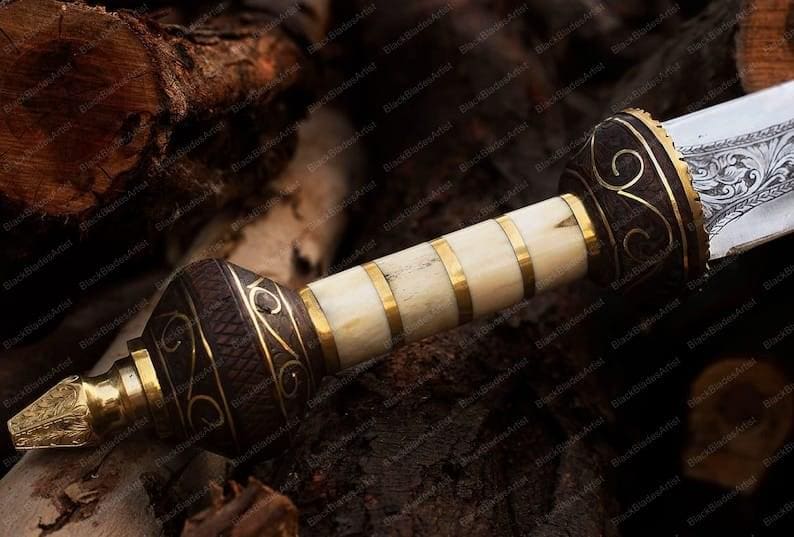 Op de afbeelding: Close-up van een decoratief mes met een gedetailleerd handvat en lemmet. Het handvat heeft een combinatie van donkerbruine, cr&egrave;mekleurige en goudkleurige metalen accenten. Het lemmet heeft een gegraveerd ontwerp. Het mes ligt op een donker houten oppervlak.