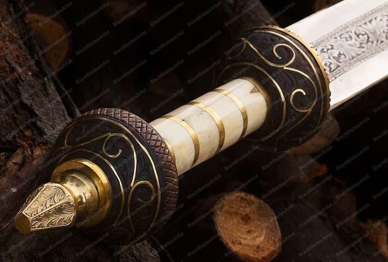 Op de afbeelding: Close-up van een decoratief zwaard met een sierlijke handgreep. De handgreep heeft een cr&egrave;mekleurige grip met gouden banden en donkerbruine accenten. De pareerstang en pommel zijn goudkleurig met ingewikkelde ontwerpen. Het lemmet is zilverkleurig met ge&euml;tste patronen.