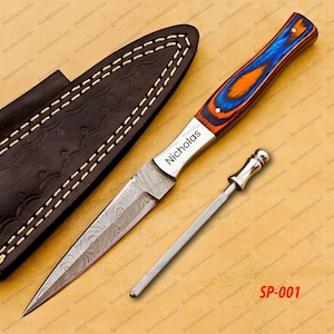 May include: A Damascus steel knife with a blue, orange, and red handle. The knife has a leather sheath and a sharpening steel. The blade is engraved with the name "Nicholas".