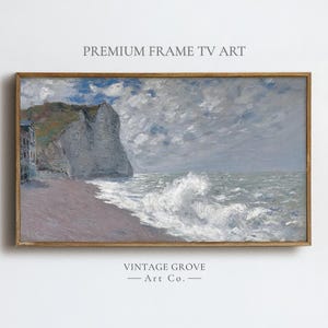 May include: A framed landscape painting of a coastal scene. The artwork depicts a cliffside, beach, and ocean with crashing waves. The sky is overcast with shades of gray and blue. The frame is a light brown color. Text on the top reads "PREMIUM FRAME TV ART".
