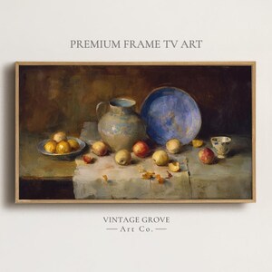 Pode incluir: Pintura de natureza morta emoldurada com um jarro de cerâmica azul e bege, um prato azul e uma pequena tigela de frutas amarelas. Maçãs e pêssegos espalhados sobre uma toalha de mesa branca. O texto "PREMIUM FRAME TV ART" e "VINTAGE GROVE - Art Co." são visíveis.