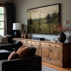 May include: A living room scene featuring a large landscape painting above a wooden cabinet. The cabinet has multiple drawers and doors. A dark gray sofa with orange and brown pillows is in the foreground. A black lamp and vases are on the cabinet.