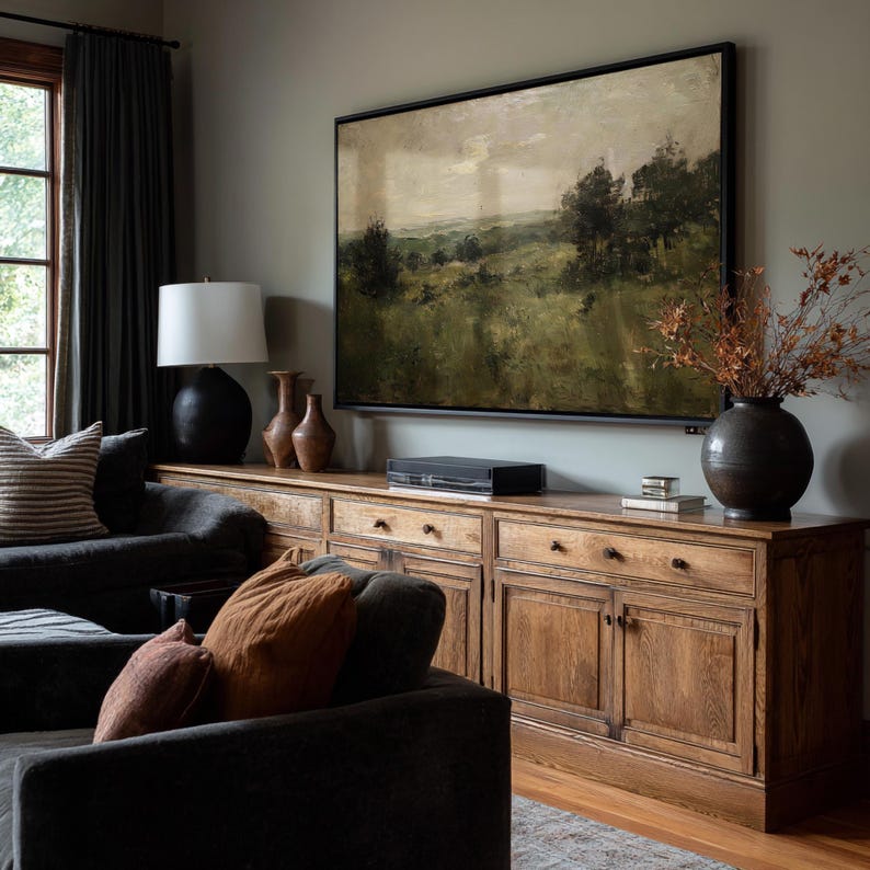 May include: A living room scene featuring a large landscape painting above a wooden cabinet. The cabinet has multiple drawers and doors. A dark gray sofa with orange and brown pillows is in the foreground. A black lamp and vases are on the cabinet.
