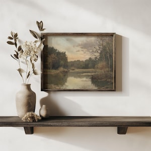 May include: A framed landscape painting depicting a lake surrounded by trees. The painting is in a wooden frame and is hanging on a white wall. A vase with dried flowers and a small vase are on a wooden shelf below the painting.