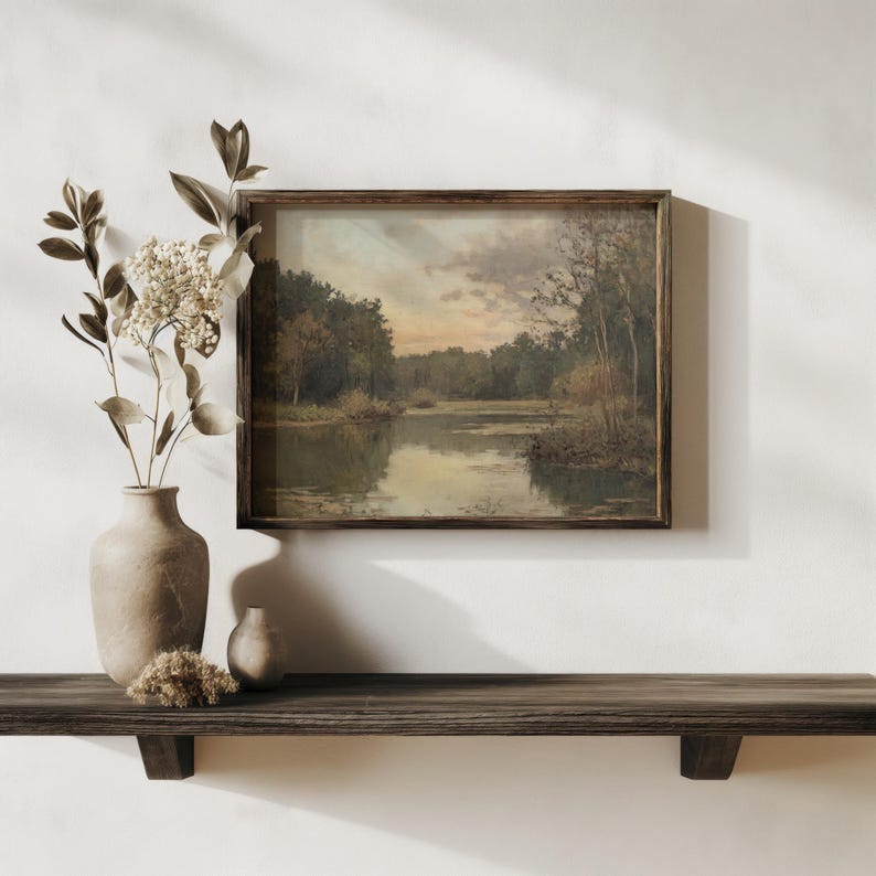 May include: A framed landscape painting depicting a lake surrounded by trees. The painting is in a wooden frame and is hanging on a white wall. A vase with dried flowers and a small vase are on a wooden shelf below the painting.