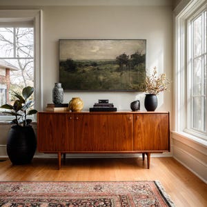 May include: A long wooden sideboard with four doors and tapered legs, topped with a landscape painting, a speckled gray vase, a yellow textured vase, a stack of books, and a black vase containing dried branches, next to a large potted plant.