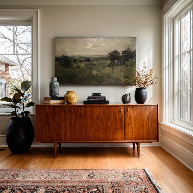 May include: A long wooden sideboard with four doors and tapered legs, topped with a landscape painting, a speckled gray vase, a yellow textured vase, a stack of books, and a black vase containing dried branches, next to a large potted plant.