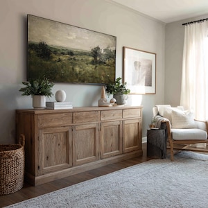 May include: A light-colored wooden cabinet with four doors and two drawers, topped with decorative items and greenery. A landscape painting hangs above the cabinet. A woven basket and a chair with a patterned rug are in the foreground.