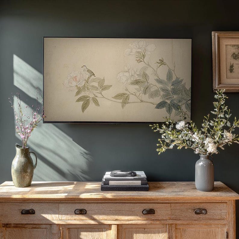 May include: A framed artwork featuring a bird perched on a flowering branch. The artwork is displayed above a wooden cabinet. A vase with flowers is on the left, and another vase with flowers is on the right. The wall is painted a dark green.