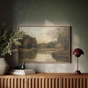 May include: A framed landscape painting depicting a lake surrounded by trees. The painting is hanging on a wall above a wooden cabinet with a red mushroom-shaped lamp on the right side.