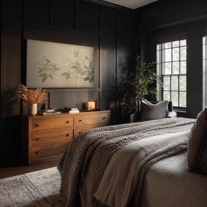 May include: A bedroom with dark walls and a wooden dresser. A large framed artwork hangs above the dresser. A bed with a gray knitted blanket and pillows is in the foreground. A vase of dried flowers and a lit candle sit on the dresser.