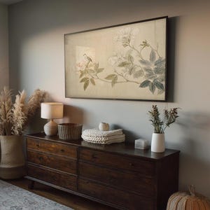 May include: A dark wood dresser with six drawers, topped with a framed botanical print, a lamp, a woven basket, and decorative items. The print features a bird perched on a flowering branch. A large vase with dried pampas grass is on the left.