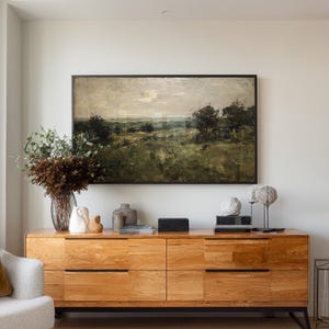 May include: A wooden dresser with six drawers, topped with decorative items and a framed landscape painting. The painting depicts a field with trees under a cloudy sky. A vase of flowers and various sculptures are on the dresser.
