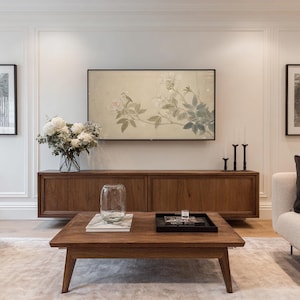 May include: A living room scene featuring a low, square wooden coffee table with a glass vase and a black tray. A wooden cabinet is below a large screen displaying floral artwork. White flowers in a glass vase and two black candle holders are also visible.