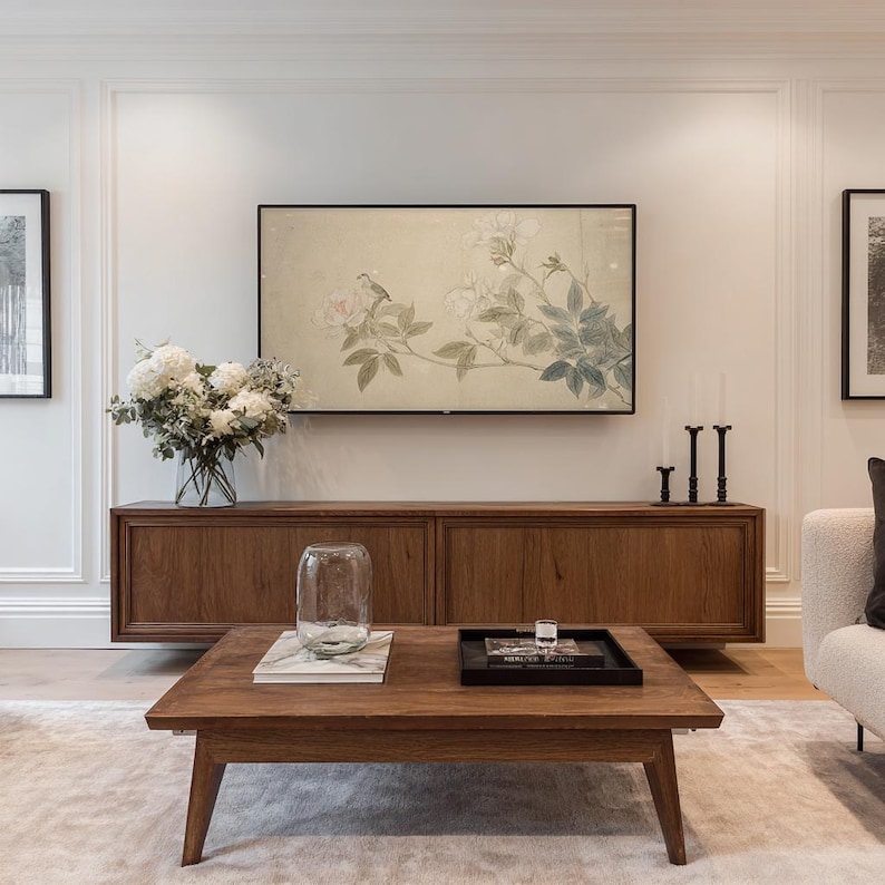 May include: A living room scene featuring a low, square wooden coffee table with a glass vase and a black tray. A wooden cabinet is below a large screen displaying floral artwork. White flowers in a glass vase and two black candle holders are also visible.