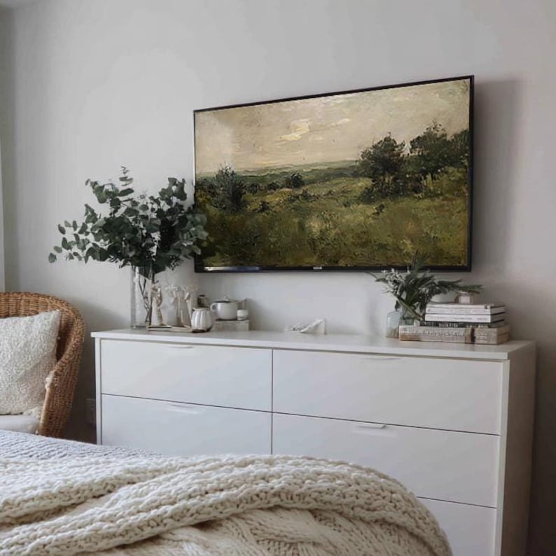 May include: A bedroom scene featuring a white dresser with six drawers, topped with a landscape painting in a black frame. A clear vase with green foliage and decorative items sit on the dresser. A woven chair and a cream-colored knitted blanket are also visible.