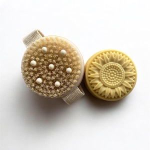 May include: A round, natural bristle body brush with a cream-colored strap and a round bar of soap with a sunflower design. The brush has white, rounded massage nodules.