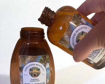 May include: Two amber-colored glass bottles of foaming hand soap. One bottle is being poured into the other. The labels read "Sunflower Beach Co. Seaside Resort" and include a palm tree and sunflower graphic. Each bottle contains 8 fl oz (236ml).