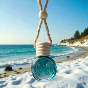 May include: A round, clear glass air freshener with a blue liquid inside, featuring a beach scene with palm trees and a sun design. It hangs from a beige rope and wooden cap. The background shows a beach and ocean.