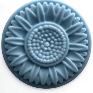 May include: A round, blue-grey soap bar with a sunflower design. The soap features a detailed embossed sunflower with petals and a textured center. The soap is a solid color and sits against a white background.