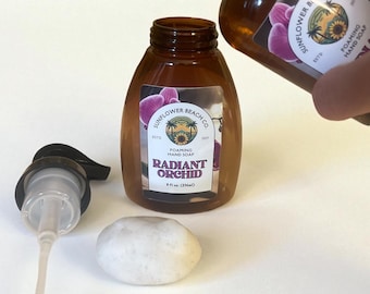 May include: Two amber-colored bottles of foaming hand soap, one being poured into the other. The bottles have labels with the text "Radiant Orchid" and "Sunflower Beach Co." A white stone and a pump dispenser are also visible.