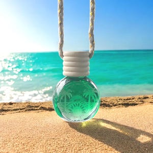 May include: A clear, spherical air freshener with a green liquid inside, featuring a palm tree design. It hangs from a white and gold rope, with a white cylindrical base. The background shows a beach and ocean.