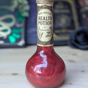 Op de afbeelding: Een klein glazen flesje met een rode vloeistof erin. De fles heeft een kurken stop met een bruin touw eromheen gewikkeld. Het etiket op de fles staat "Magie & Mixturen Health Potion - by DeepCosmicBytes".