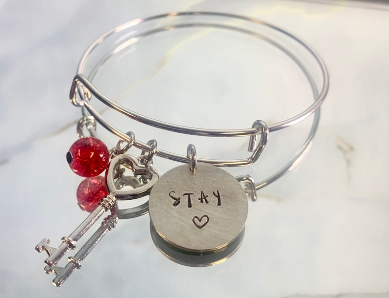 以下が含まれることがあります： ハートのチャーム、キーのチャーム、そして「STAY」とハートが書かれた丸いチャームが付いたシルバーのチャームブレスレット。ブレスレットには、赤いグリッタービーズも2つ付いています。