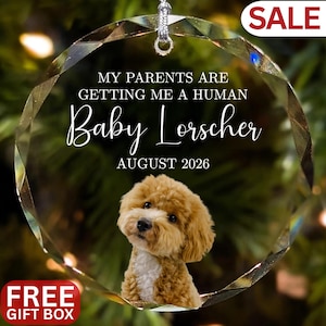 May include: A clear, faceted glass ornament with a silver loop for hanging. The ornament features a fluffy, brown and white dog and text that reads "MY PARENTS ARE GETTING ME A HUMAN Baby Lorscher AUGUST 2026". A "SALE" and "FREE GIFT BOX" tag are also visible.