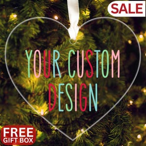 Op de afbeelding: Transparante hartvormige kerstversiering met de tekst "YOUR CUSTOM DESIGN" in kleurrijke letters. Een wit lint is bevestigd om op te hangen. De versiering staat tegen een achtergrond van een kerstboom met lichtjes. De woorden "SALE" en "FREE GIFT BOX" zijn ook zichtbaar.