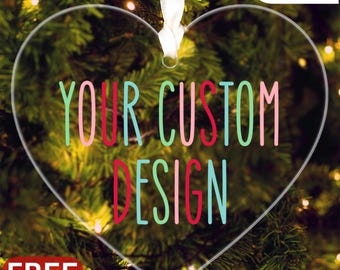 Puede incluir: Adorno transparente en forma de corazón con el texto "YOUR CUSTOM DESIGN" en letras coloridas. Se adjunta una cinta blanca para colgarlo. El adorno está sobre un fondo de árbol de Navidad con luces. También se ven las palabras "SALE" y "FREE GIFT BOX".
