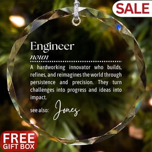 May include: A clear, faceted glass ornament with the definition of "Engineer" printed in black. The definition describes an innovator. The ornament has a silver string for hanging and the word "SALE" is in the upper right corner.