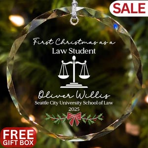 May include: A clear, faceted glass Christmas ornament with the text "First Christmas as a Law Student" and the name "Oliver Willis". Includes a scale of justice graphic, holly, and a red bow. The ornament is from Seattle City University School of Law, 2025.
