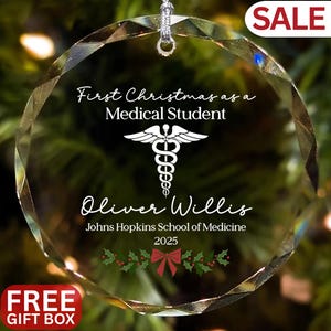 May include: A clear, faceted glass Christmas ornament with the text "First Christmas as a Medical Student" and a medical symbol. The ornament also includes the name "Oliver Willis", school name, and the year 2025. A red bow and holly are at the bottom.