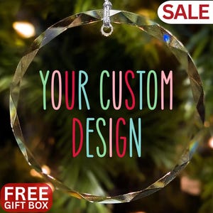 Op de afbeelding: Een heldere, facetgeslepen glazen kerstbal met de tekst "YOUR CUSTOM DESIGN" in roze, groen en blauw. De kerstbal hangt aan een zilveren draad. Het woord "SALE" staat in een rode doos en "FREE GIFT BOX" staat in een rode doos.