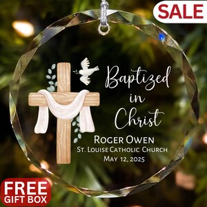 Op de afbeelding: Een heldere, facetgeslepen glazen ornament met een zilveren ophanglus. Het ornament heeft een houten kruis met een witte doek, een duif en de woorden "Baptized in Christ". Tekst bevat een naam, kerk en datum. Een "SALE" en "FREE GIFT BOX" label is zichtbaar.