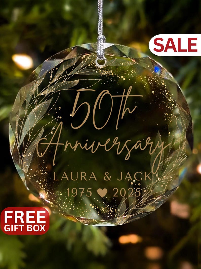 May include: A clear, faceted glass ornament with the words "50th Anniversary" in gold script. The names "Laura & Jack" and the years "1975 2025" are also inscribed. The ornament has a silver string for hanging and is decorated with gold leaf accents.