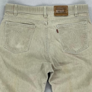 May include: Close-up of the back of tan-colored Levi Strauss & Co. jeans. The jeans have two back pockets with decorative stitching, a small red tag, and a brown leather patch with the Levi Strauss & Co. logo.