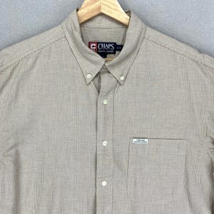 May include: A button-down shirt with a small checkered pattern in tan and white. The shirt has a collar, a chest pocket, and the "CHAPS RALPH LAUREN" logo. The shirt is hanging on a wooden hanger.