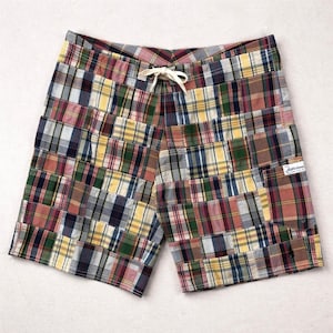 May include: Multi-colored plaid patchwork shorts with a drawstring waist. The shorts feature a variety of plaid patterns in shades of red, blue, yellow, and green. A small label is sewn on the right side. The shorts are made of a lightweight fabric.