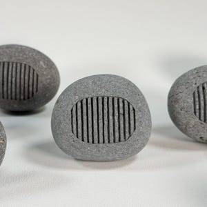 May include: Several gray, oval-shaped stones with a carved, rectangular pattern of vertical lines. The stones are arranged on a white surface, with a few in focus and others blurred in the background. The stones appear to be natural and have a smooth texture.