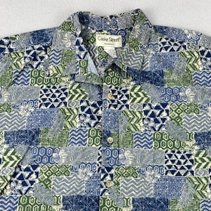 May include: A button-down shirt with a patchwork design in shades of blue, green, and white. The shirt features a collared neckline and the "Cooke Street Honolulu" label. The pattern includes geometric shapes and floral motifs.
